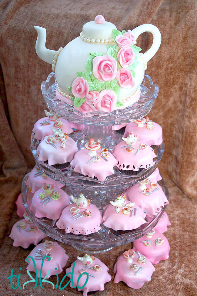 Teapot cake decorated with fondant roses and tea party cupcakes on a tiered glass cake stand.