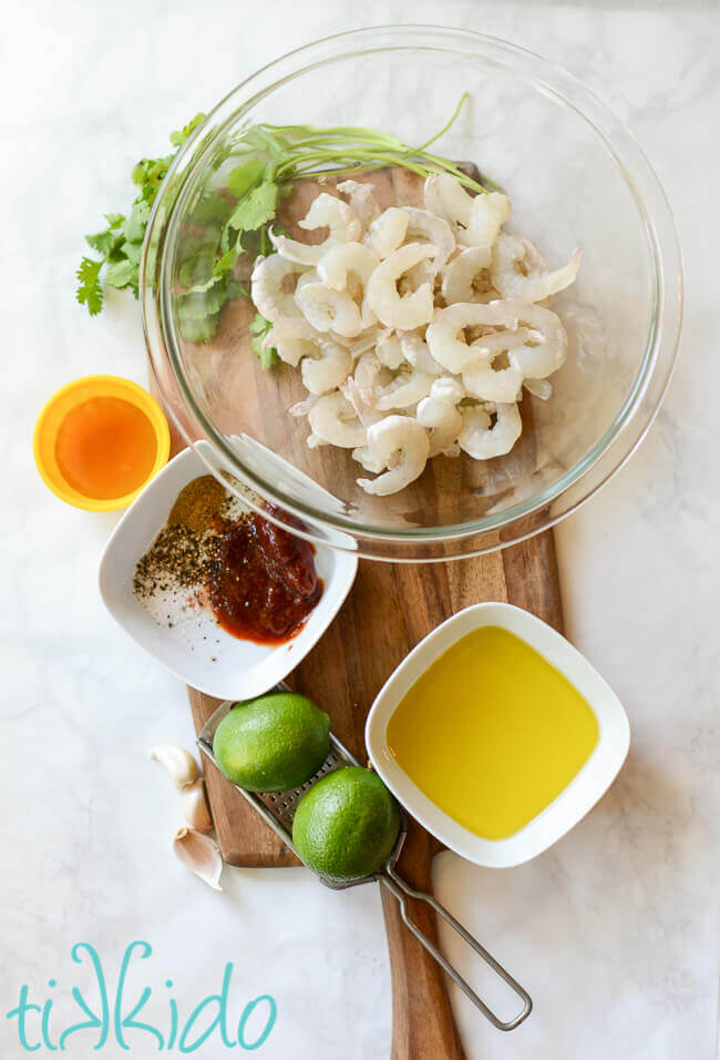 Ingredients for tequila lime grilled shrimp on a wooden cutting board on a white marble surface.