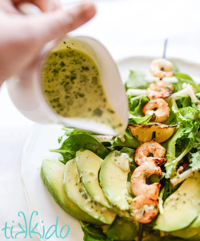 Homemade tequila lime salad dressing being poured on a salad topped with a skewer of grilled shrimp.
