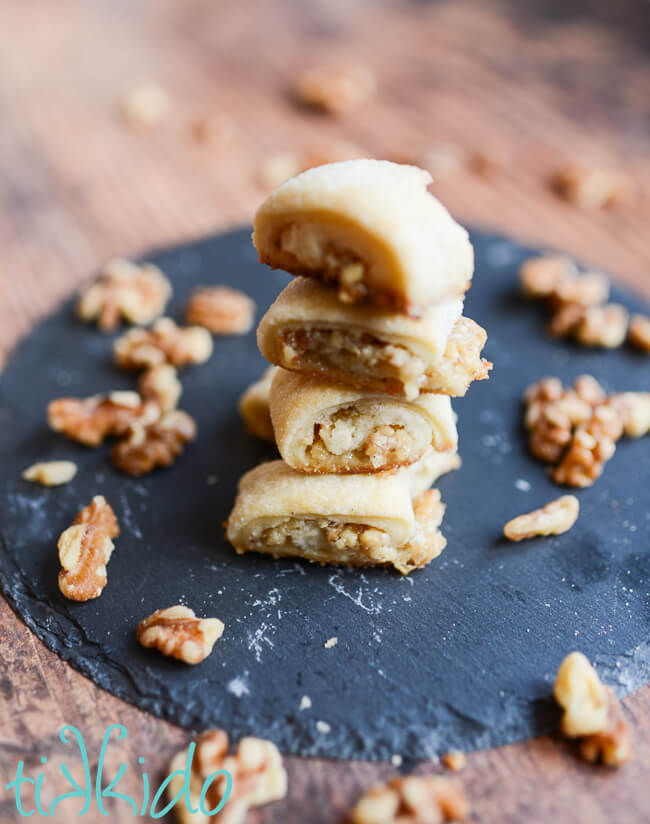 Stack of walnut rugelach cookies on a round piece of slate, with scattered walnut pieces surrounding the cookies.