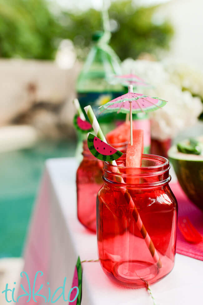 Pink mason jars for drinks, garnished with wedges of watermelon and watermelon themed cocktail umbrellas and straws.