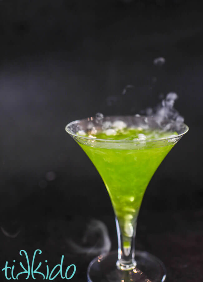 Side shot of a green, shimmery Halloween cocktail bubbling with dry ice smoke.