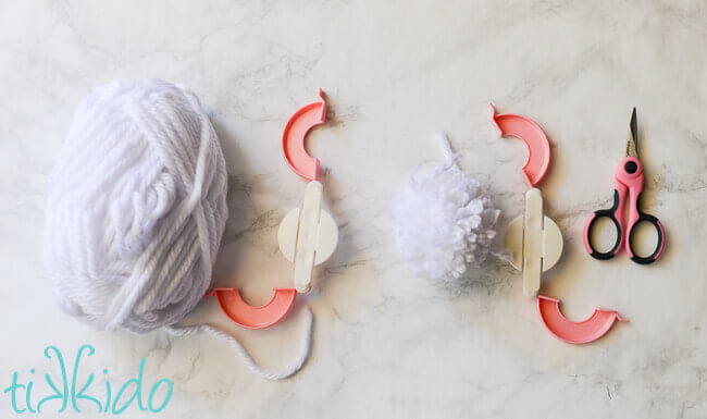 White yarn being turned into a white pom pom with a pink pom pom making tool.