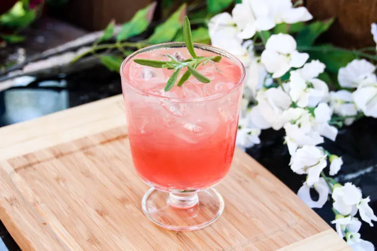 Glass of summer buffalo pink cocktail on a wooden cutting board, with white flowers in the background.