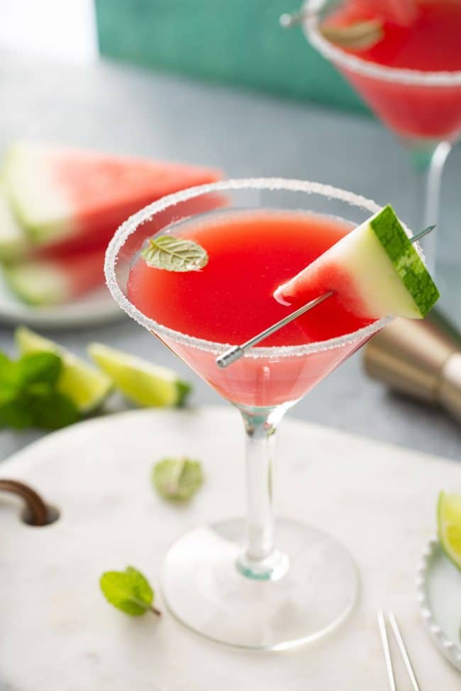 Deep pink watermelon martini served in a classic martini glass rimmed with sugar, and garnished with a triangular slice of watermelon and a fresh mint leaf.