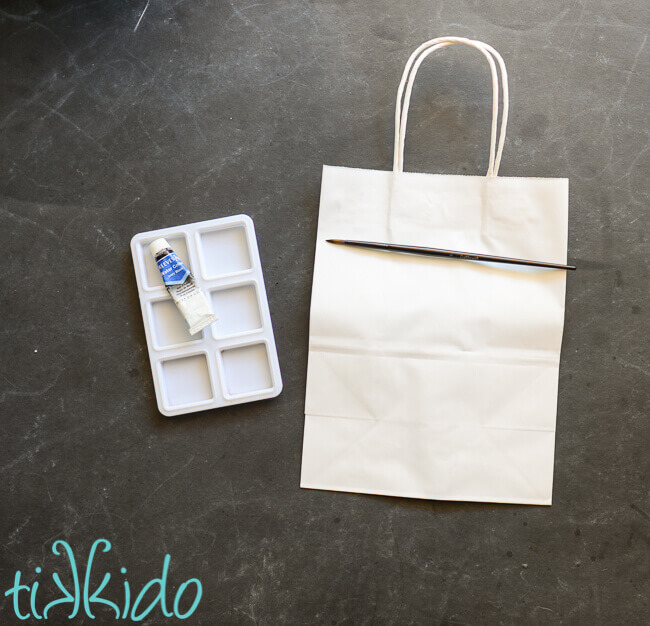 Paint brush on top of plain white gift bag, next to a small paint tray and tube of black watercolor paint.