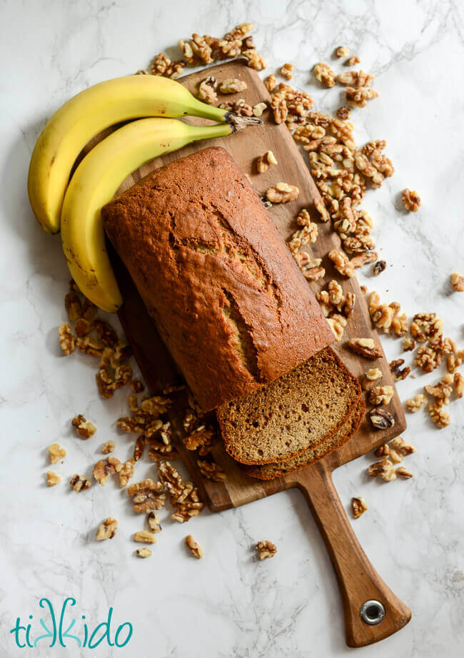 Loaf of banana bread on a cutting board, surrounded by bananas and walnuts.