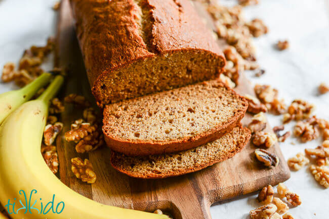 Loaf of banana bread, sliced on a cutting board, surrounded by walnuts and bananas.