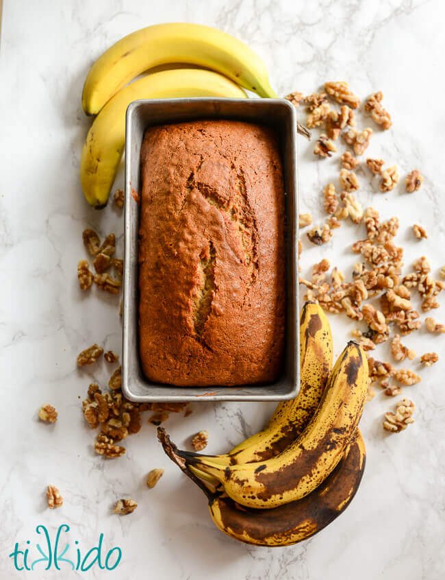 Loaf of banana bread in pan, surrounded by bananas and walnuts.
