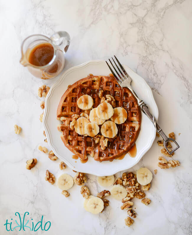 Banana bread waffles topped with sliced bananas, walnuts, and brown sugar caramel syrup on a white plate with a fork, small pitcher of syrup above the plate, and scattered banana slices and walnuts around the plate.