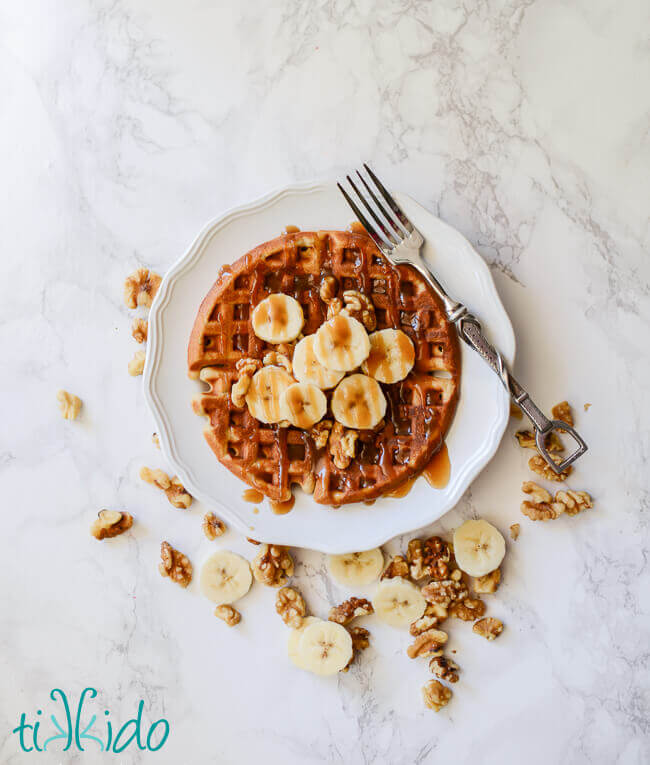 Banana bread waffles topped with sliced bananas, walnuts, and brown sugar caramel syrup on a white plate with a fork, small pitcher of syrup above the plate, and scattered banana slices and walnuts around the plate.