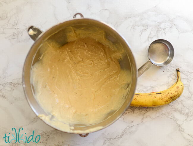 Mixer bowl with banana bread batter, banana and 1/2 cup measure on the white marble surface beside the bowl.