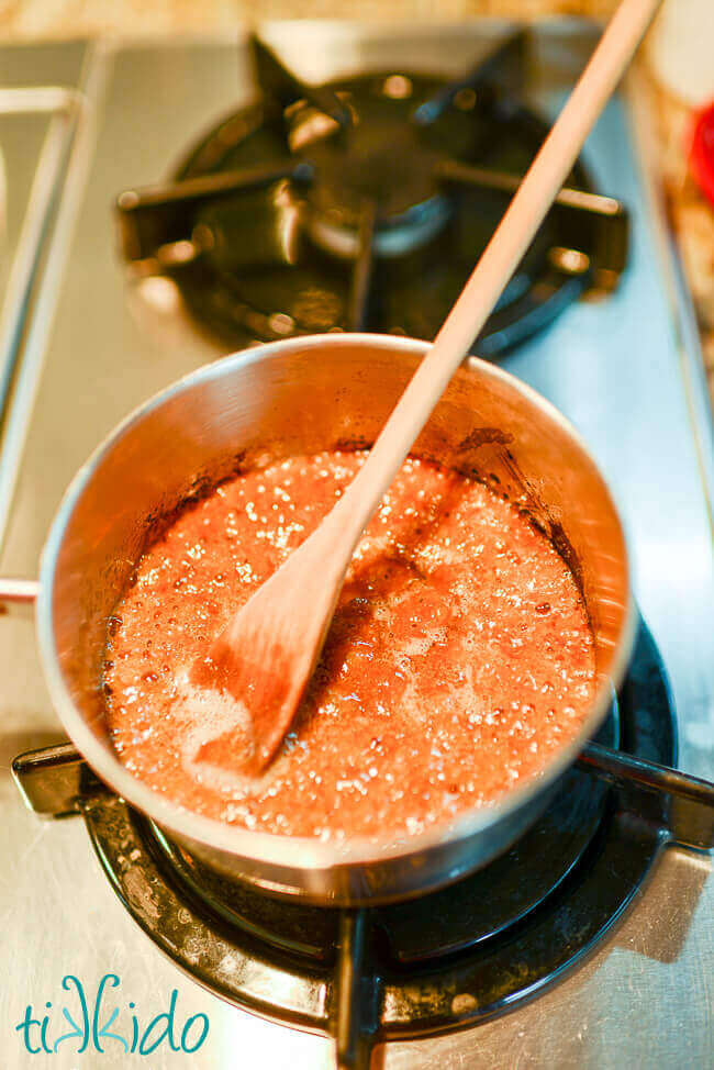 Small saucepan with bubbling brown sugar caramel syrup being cooked inside, wooden spoon in pot.  On a gas burner stove.