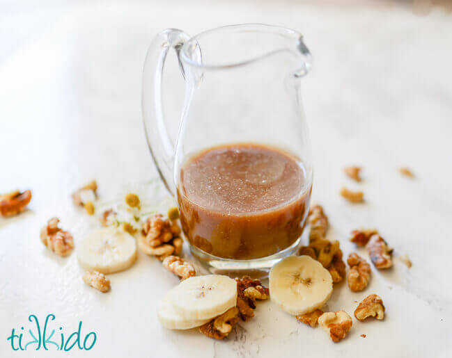 Small pitcher of brown sugar caramel syrup on a white marble background with sliced bananas and walnuts around the base.