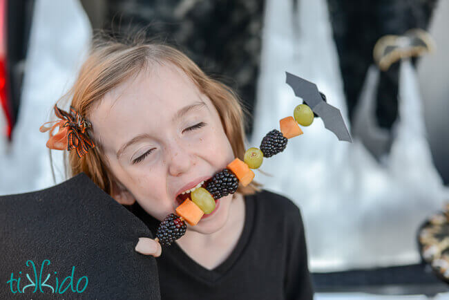 Little girl eating healthy Halloween fruit skewer topped with a cute little bat.