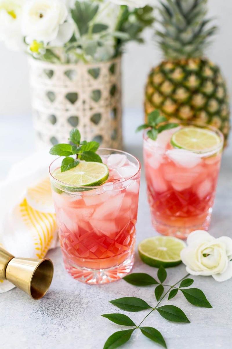 Two glasses with pink bay breeze cocktails garnished with a slice of lime and fresh mint, in front of a fresh pineapple and a vase of flowers.