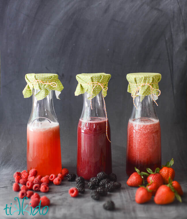 Raspberry simple syrup, blackberry simple syrup, and strawberry simple syrup in three carafes on a black chalkboard background.