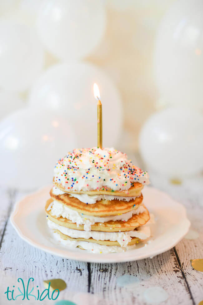 Stack of Birthday Cake Pancakes with whipped cream and sprinkles, topped with a single lit candle.