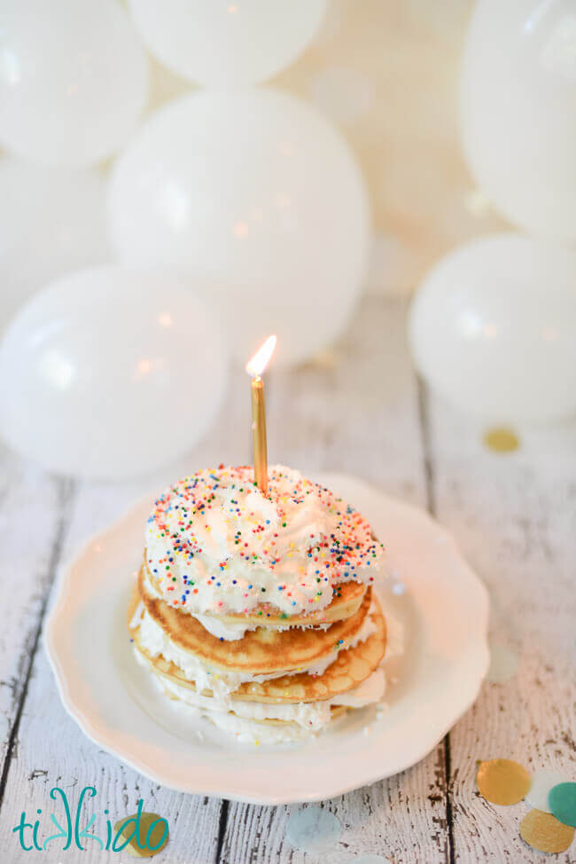 Stack of Birthday Cake Pancakes with whipped cream and sprinkles, topped with a single lit candle.