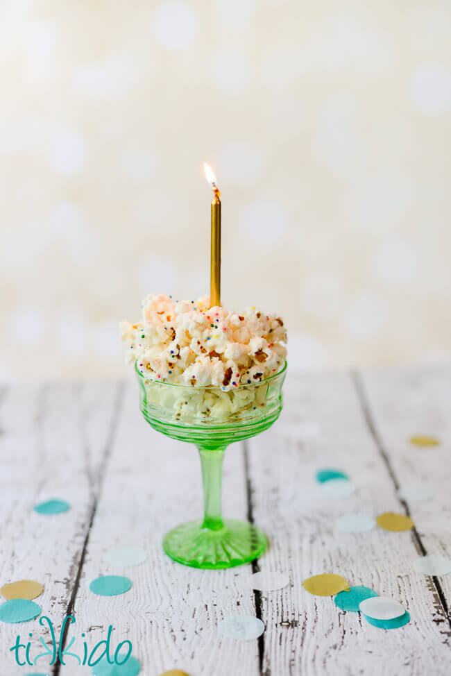 Birthday cake popcorn in a green glass dish, topped with a gold birthday candle.