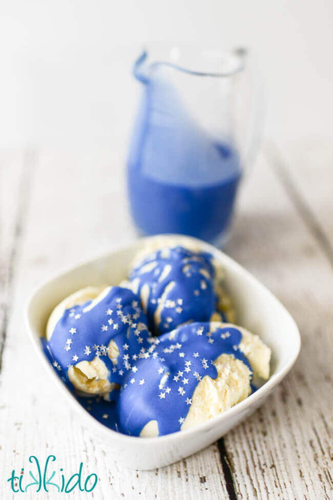 Blue Magic Shell Ice Cream Topping over vanilla ice cream sprinkled with silver star sprinkles.  A small pitcher of the Blue Magic Shell Ice Cream Topping sits in the background.