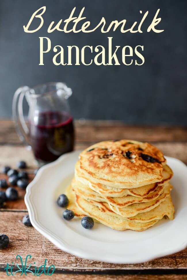 Stack of homemade buttermilk pancakes with blueberries on a white plate, next to a small pitcher of blueberry syrup.