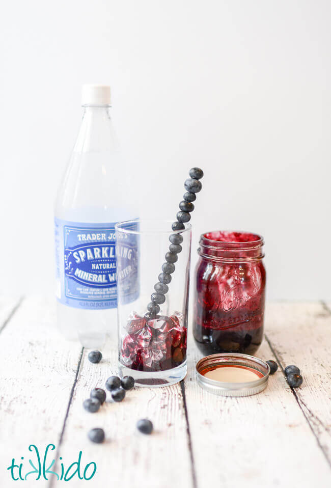 Bottle of sparkling water, jar of homemade blueberry syrup, and glass with ice, blueberry syrup, and skewered blueberries drink stirrer.