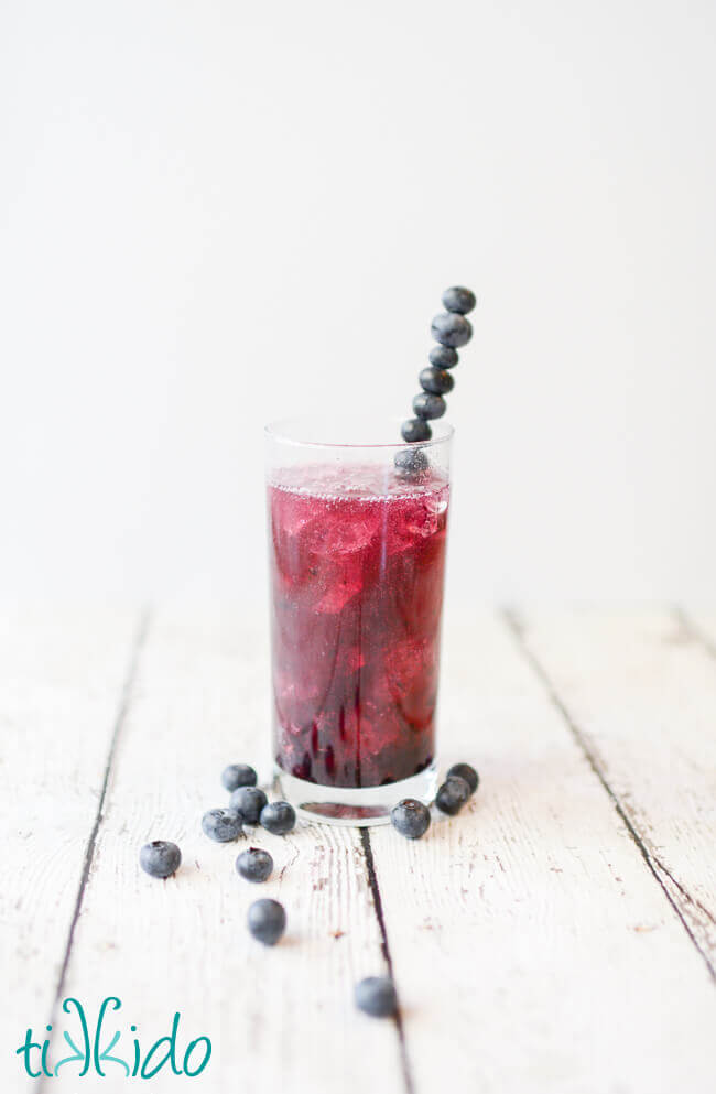 Tall glass of a vibrant bluish-purple blueberry soda with ice, with a stirrer made from skewered blueberries, on a white surface with fresh blueberries scattered around.