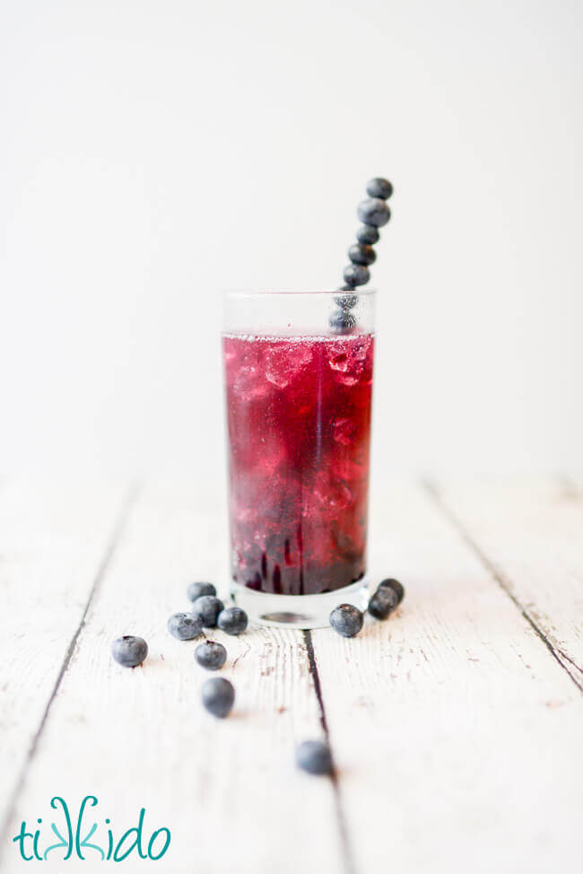 Tall glass of a vibrant bluish-purple blueberry soda with ice, with a stirrer made from skewered blueberries, on a white surface with fresh blueberries scattered around.