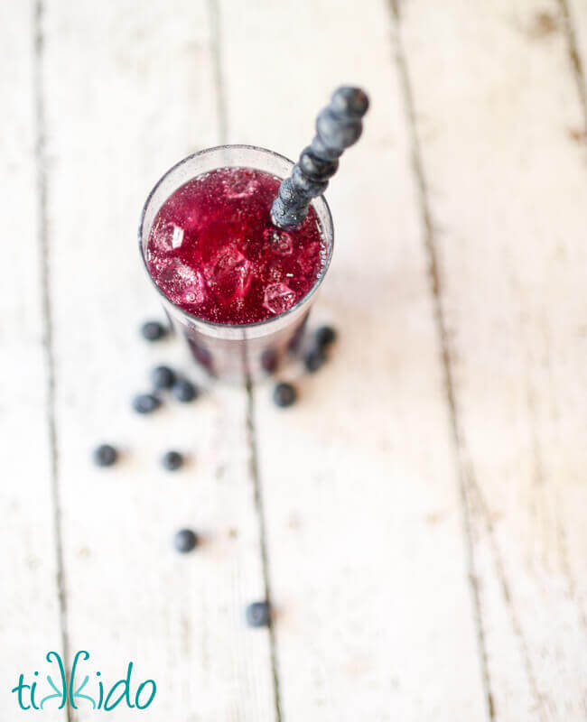 Top view of a tall glass of a vibrant bluish-purple blueberry soda with ice, with a stirrer made from skewered blueberries, on a white surface with fresh blueberries scattered around.