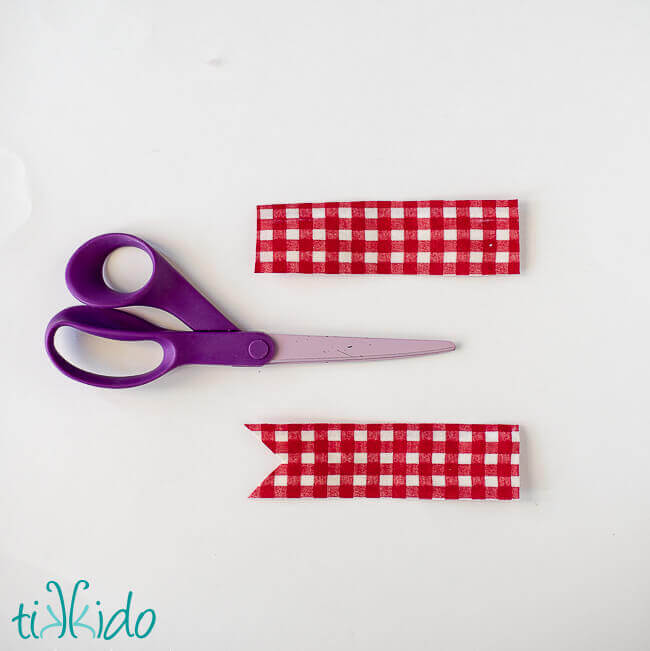 Red and white checked fabric being cut into pennant flag shapes for DIY waving flag cake toppers.