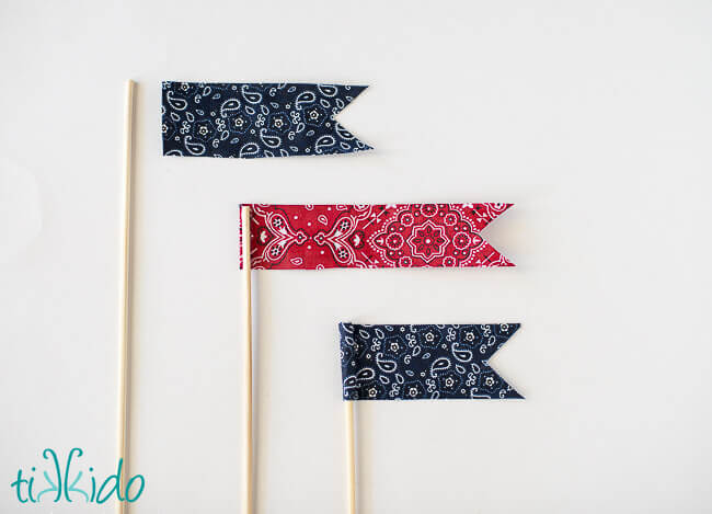 Three fabric cake topper flags being glued to the tops of bamboo skewers.