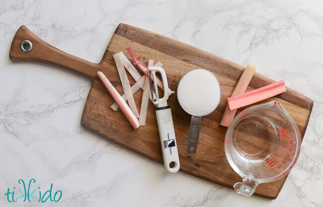 Thinly sliced rhubarb, sugar, and water, on a wooden cutting board for making candied rhubarb