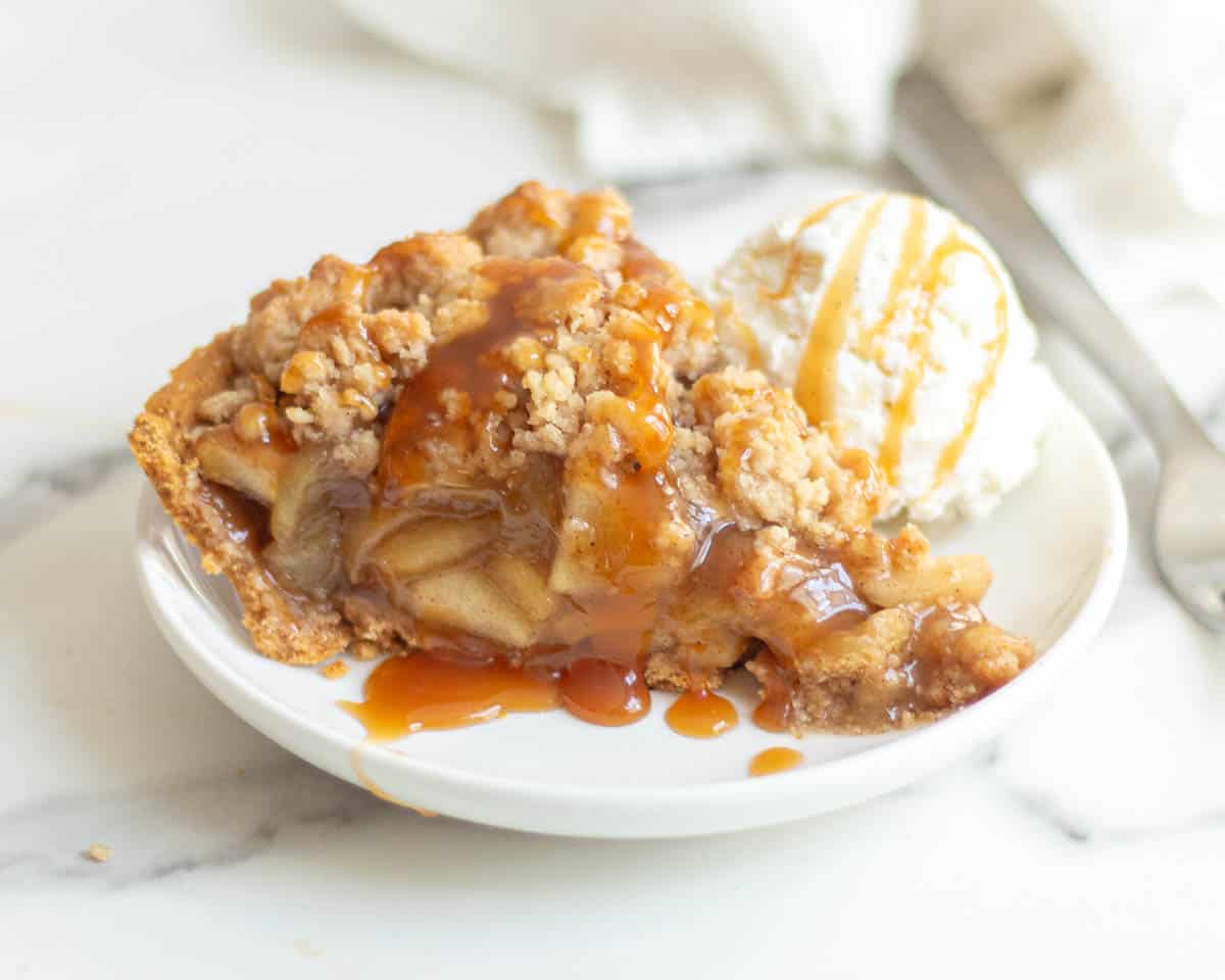 Slice of apple pie with graham cracker crust and a crumble top sitting on a white plate.  A scoop of vanilla ice cream sits next to the piece of pie, and the entire plate is drizzled with caramel syrup.