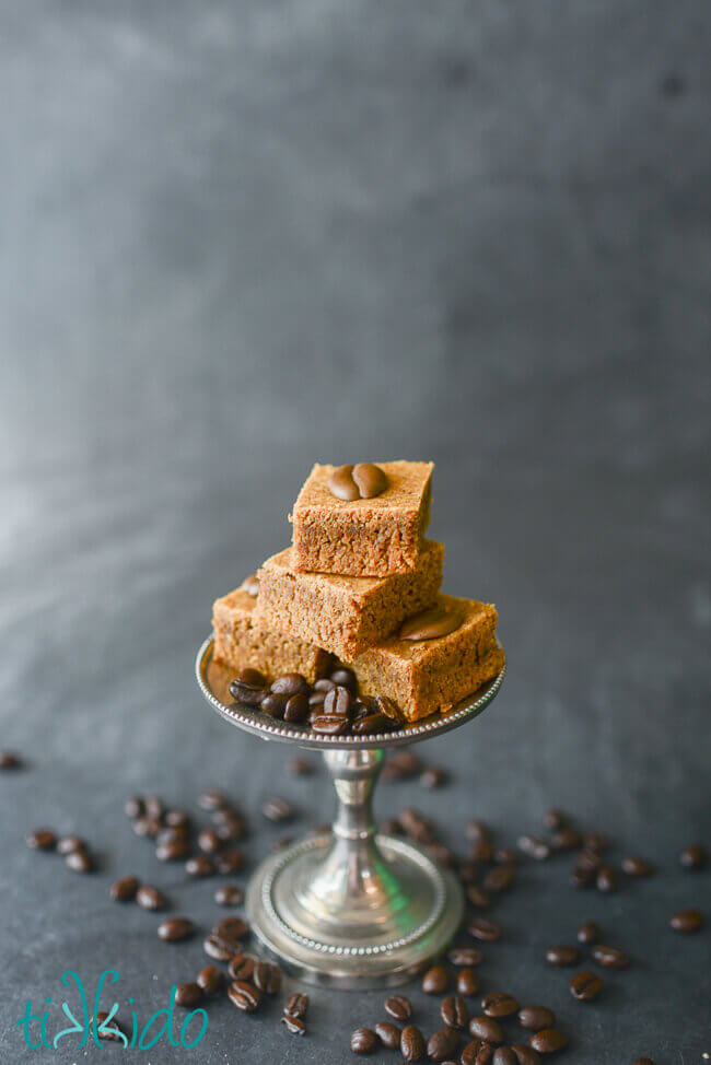 Stack of four cappuccino blondies decorated with royal icing coffee beans, on a small silver cake stand, surrounded by whole coffee beans.