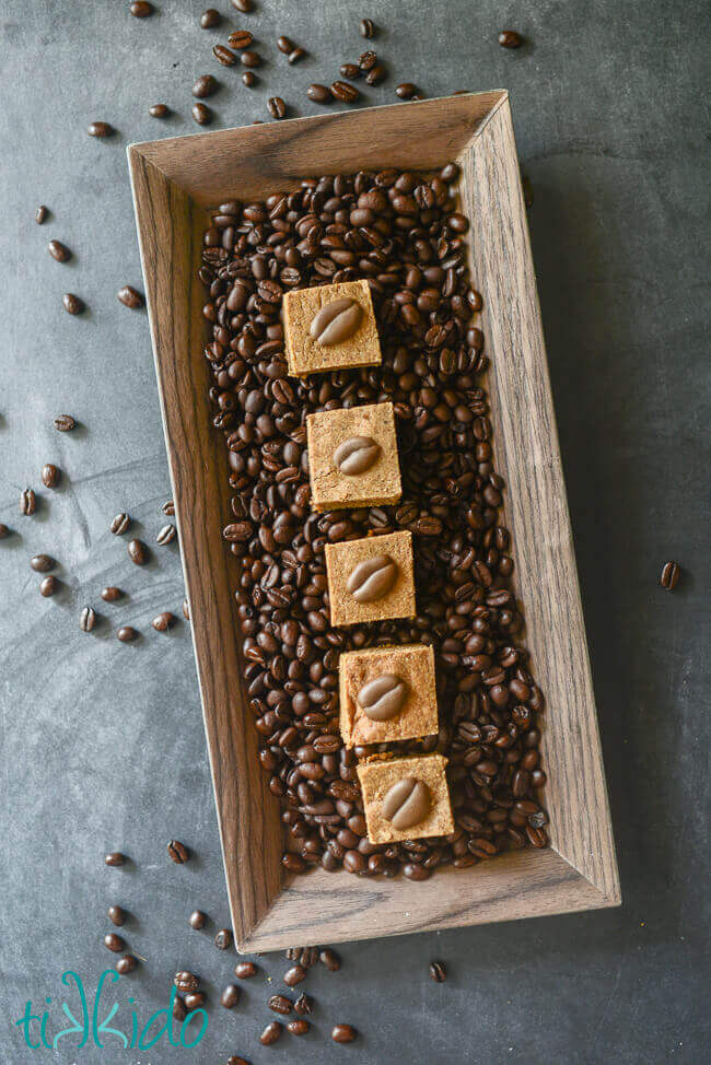 Stack of four cappuccino blondies decorated with royal icing coffee beans, arranged on a rectangular wooden tray, surrounded by whole coffee beans.