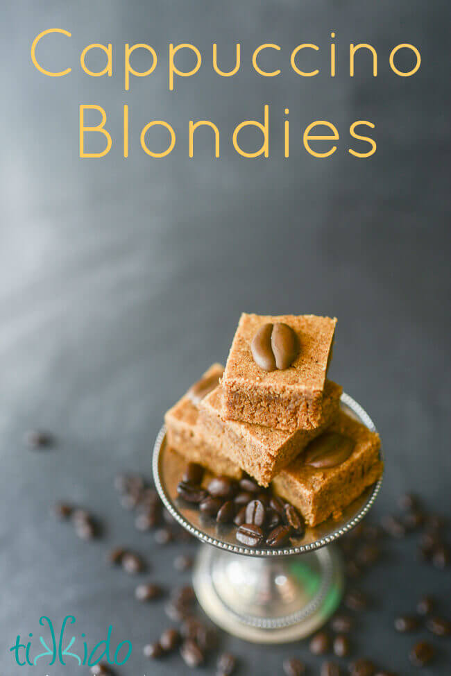 Stack of four cappuccino blondies decorated with royal icing coffee beans, on a small silver cake stand, surrounded by whole coffee beans.  Text overlay in yellow letters reads "Cappuccino Blondies."