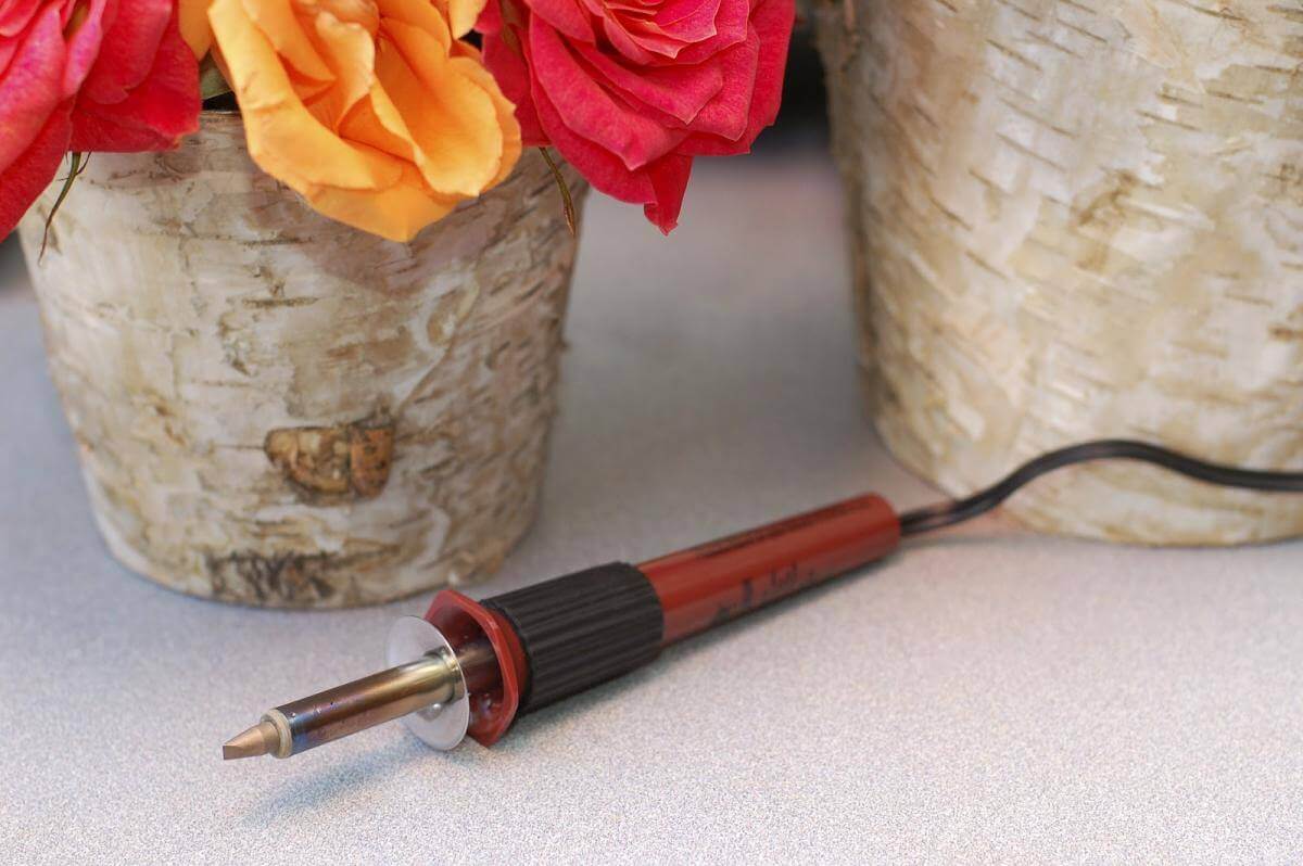 Two birch bark covered zinc floral containers with a wood burning tool on a counter.