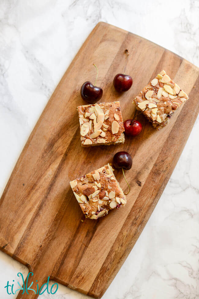 Three Cherry Almond Bar Cookies on a wooden cutting board, next to four fresh cherries.