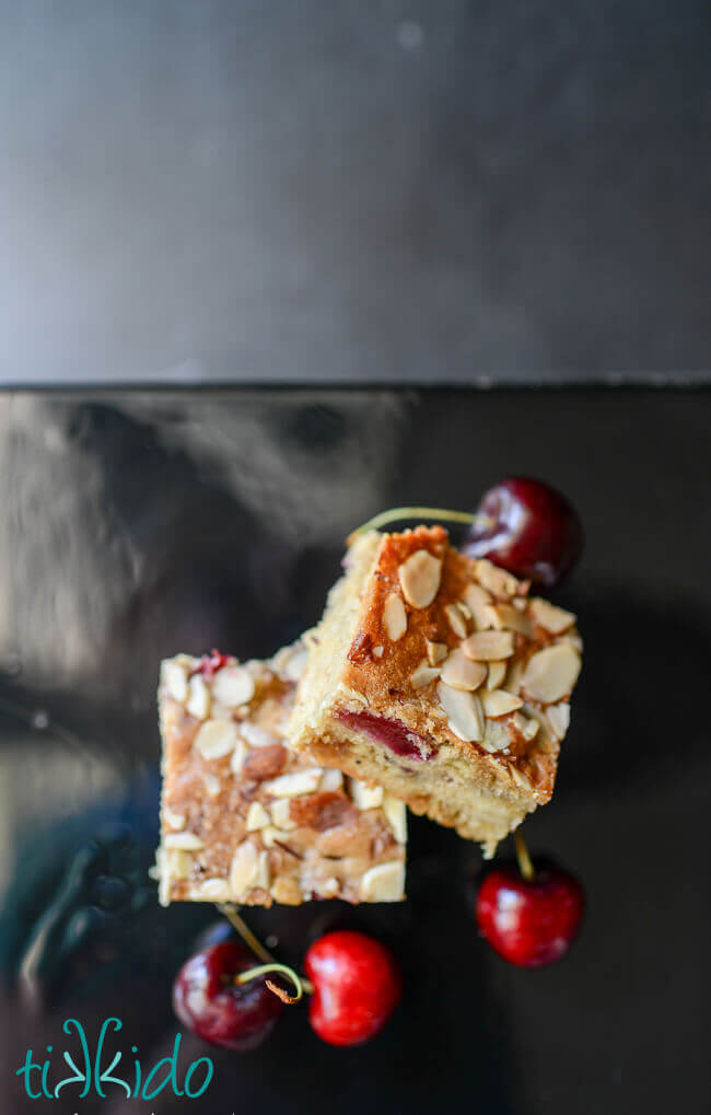Two Cherry Almond Bar Cookies on a black cake stand, surrounded by fresh cherries.