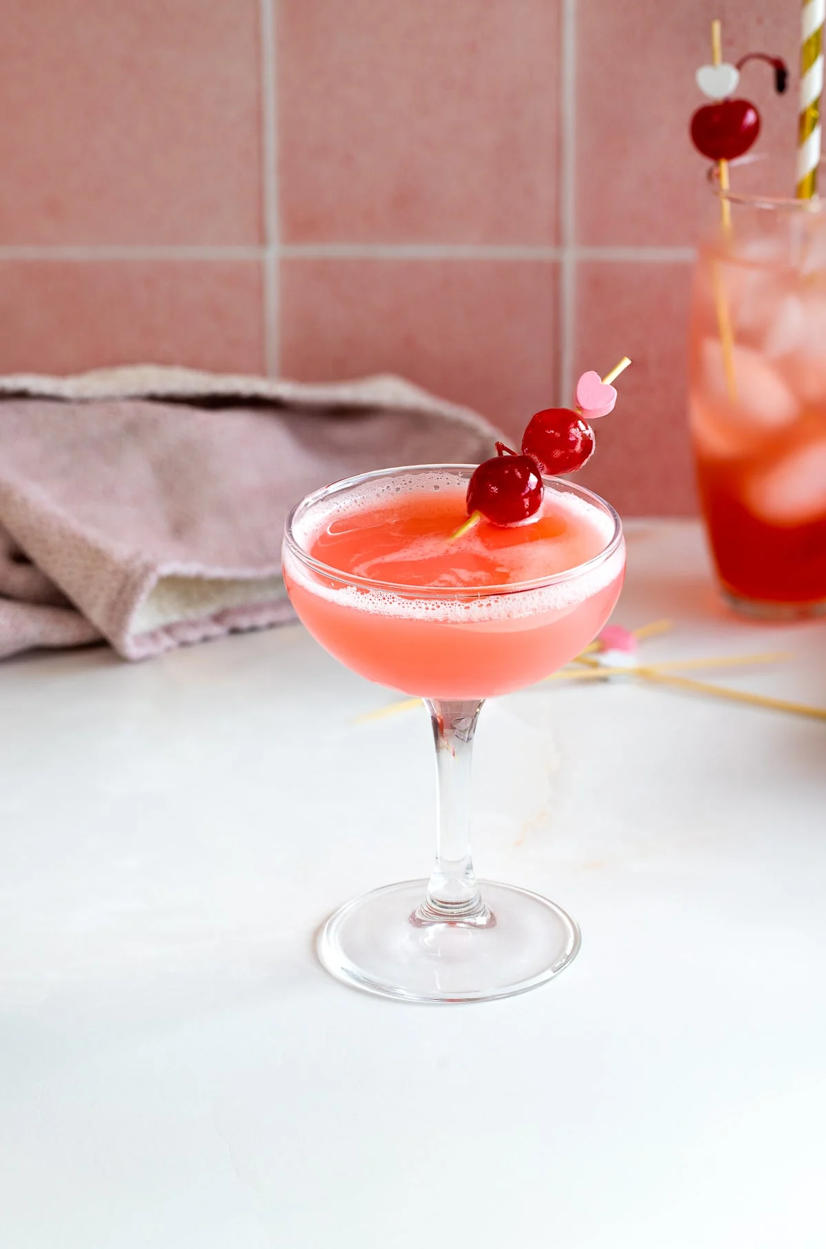 Old fashioned champagne glass filled with a pink cocktail, a Cherry Vodka Sour, garnished with a stick skewering two maraschino cherries.