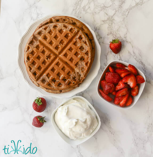 Freshly baked chocolate waffles on a white plate next to bowls of strawberries and whipped cream