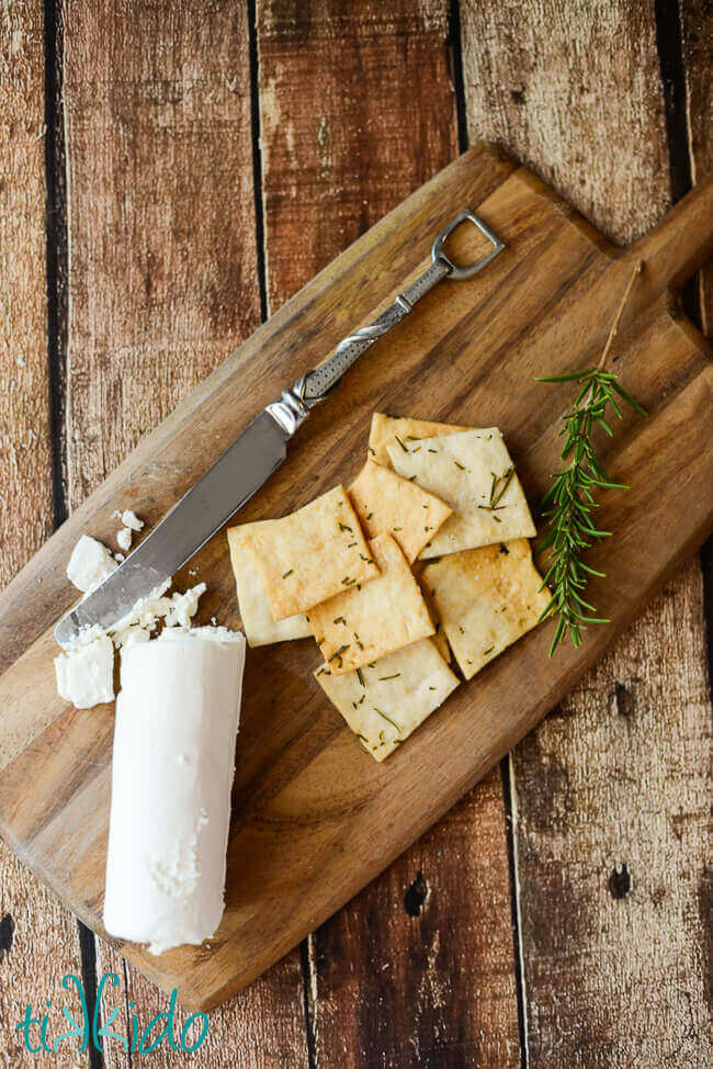 Homemade crackers with rosemary and salt on a wooden cutting board next to goat cheese.