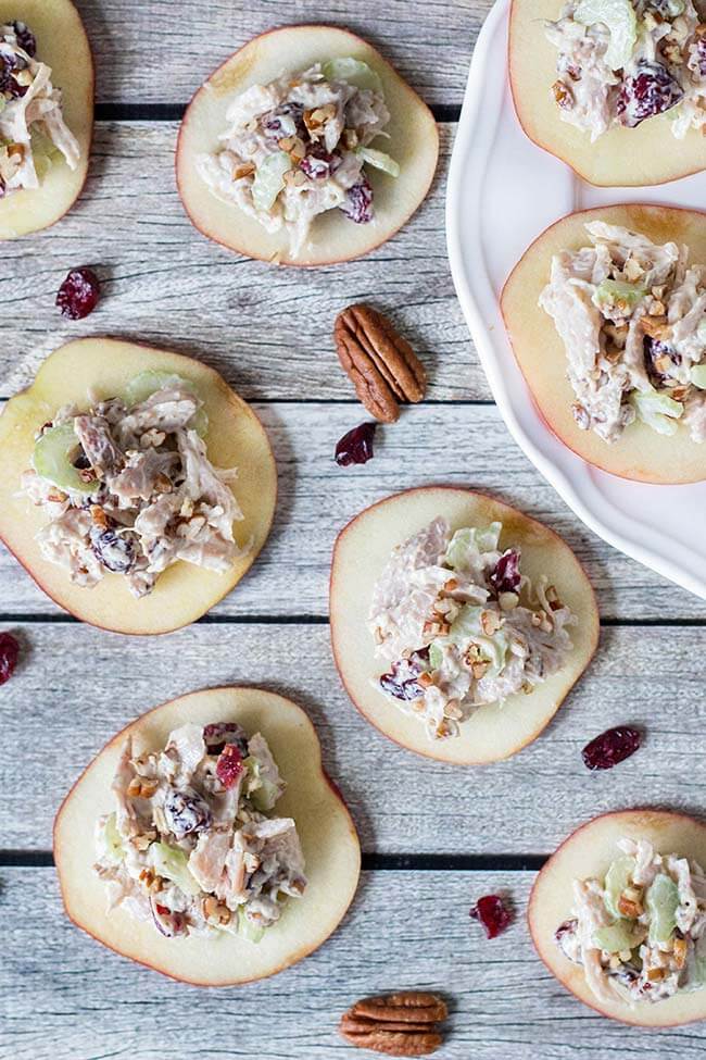 Cranberry chicken salad served on slices of apples on a light grey wooden surface.