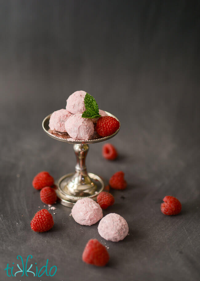 Raspberry chocolate truffles dusted with raspberry powdered sugar, surrounded by fresh raspberries on a black chalkboard background.