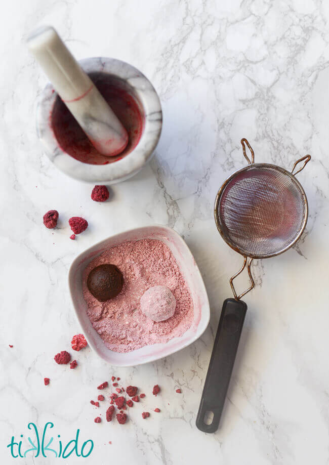 Mortar and pestle, fine mesh strainer, and bowl with raspberry powdered sugar and two truffles being rolled in the sugar. 