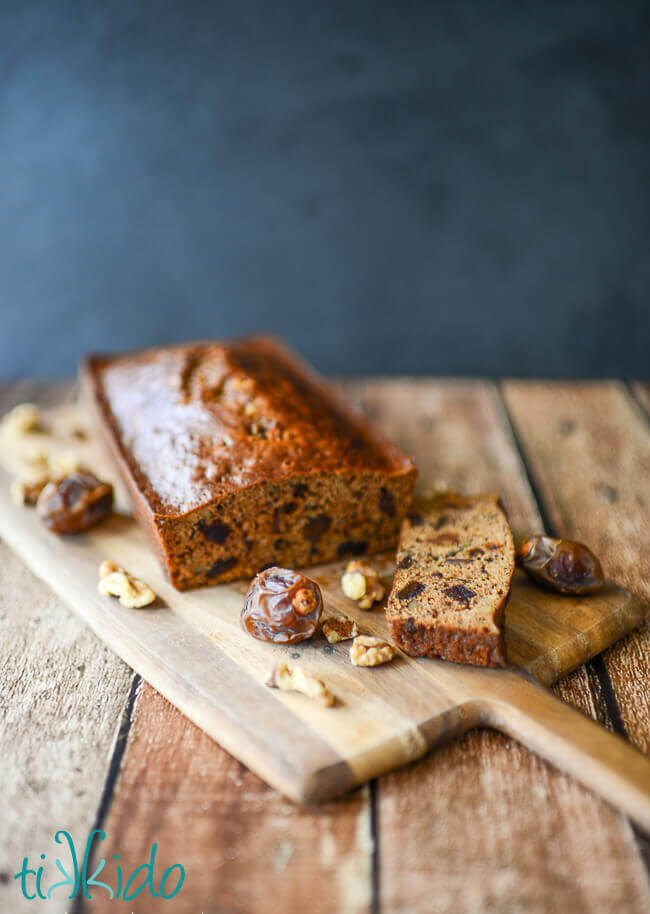 Date nut bread surrounded by dates and walnuts on a wooden cutting board.
