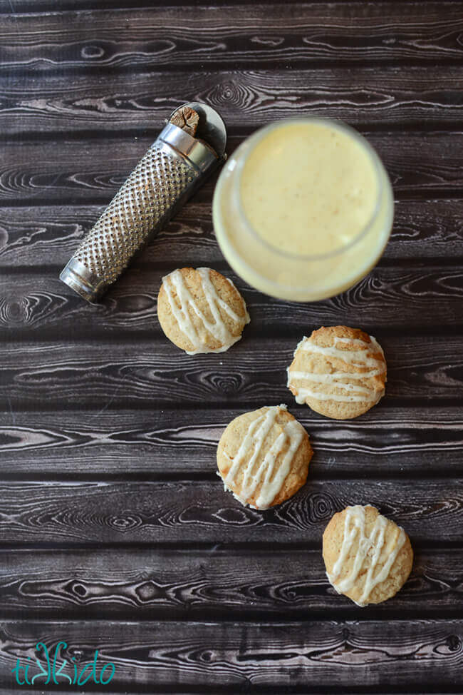 Four eggnog cookies topped with eggnog glaze on a wooden table, next to a glass of eggnog and a nutmeg grater.