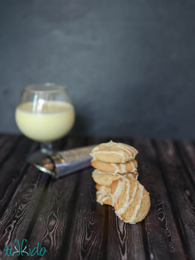 Stack of eggnog cookies in front of a glass of eggnog against a dark backdrop.