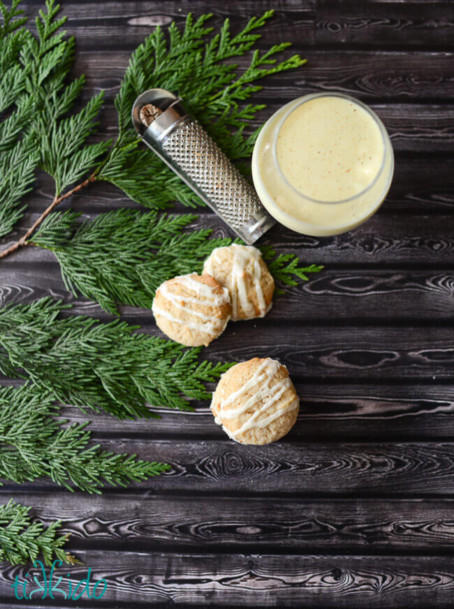 Three eggnog cookies next to a glass of eggnog and a nutmeg grater on a dark wood surface.